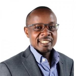 Headshot of DGH faculty Kenneth Mugwanya wearing a grey blazer, a blue colored shirt, and glasses.